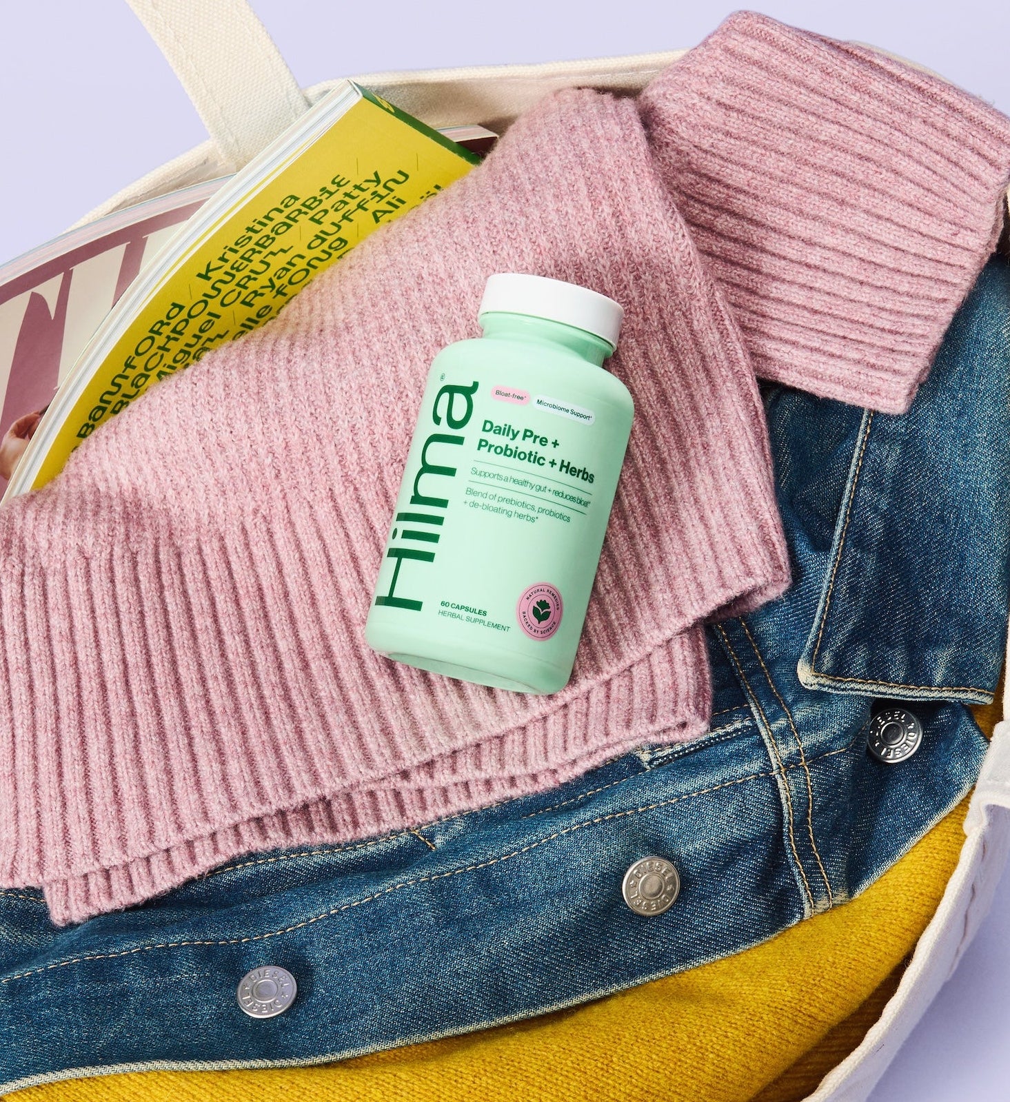 Bag with pink sweater, denim jacket, yellow shirt, and Hilma probiotic bottle on a light purple background
new