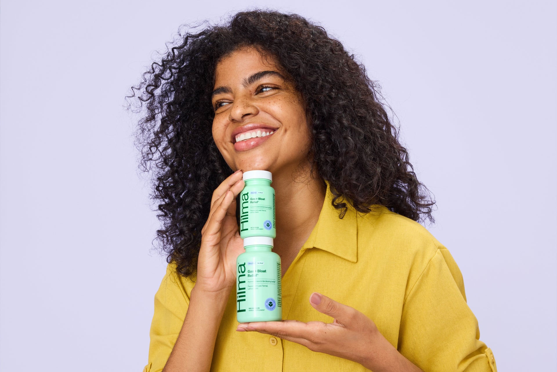 Woman holding a stack of Hilma Gas + Bloat products against a light purple background

