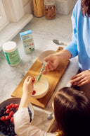 Person feeding a child with a spoon in a kitchen setting, with Huma product visible.