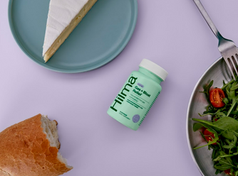 Gas + Bloat Relief bottle flatlay next to baguette, salad, and a wedge of brie cheese on a purple background
